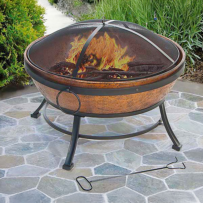 A copper fire pit with a mesh cover sits on a stone patio, surrounded by green grass. A fire is burning inside, with a metal poker on the ground nearby.